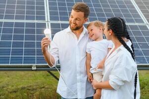 solar energía concepto. un joven, contento familia es en pie cerca solar paneles y participación un eléctrico ligero bulbo. foto