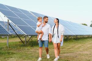 joven familia de Tres es agachado cerca fotovoltaica solar panel, pequeño chico y padres. moderno familia concepto. el concepto de verde energía foto