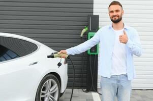 Beautiful young stylish man is with electric car at daytime charging the vehicle photo