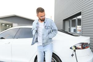 Beautiful young stylish man is with electric car at daytime charging the vehicle photo