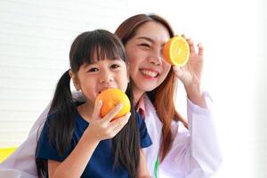 nutrición para edad escolar niños. un hermosa nutricionista o médico con un pequeño niña participación un naranja. elegir el Derecha comida según a el años de el niño para bueno salud foto