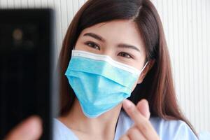 asiático hembra paciente vistiendo máscara en hospital paciente habitación ella sostiene un teléfono inteligente en línea hablando a su familia. concepto de médico servicios. pacientes infectado con coronavirus foto