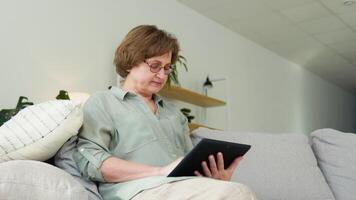 Smiling elderly woman resting on sofa, using digital tablet at home video