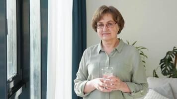 Beautiful old lady looking in camera standing in the living room of her house with a glass of water video