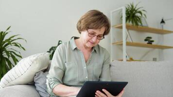 Smiling elderly woman resting on sofa, using digital tablet at home video