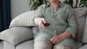 envelhecido mulher assistindo televisão dentro vivo quarto sentado em sofá segurando controlo remoto ao controle mudando televisão canais video