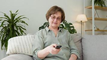 Aged woman watching tv in living room sitting on sofa holding remote control changing tv channels video