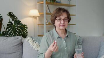 lindo Senior mulher com uma comprimido e uma vidro do água dentro dela mãos senta em uma sofá às casa video