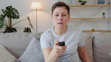 anziano signora Guardando tv nel vivente camera seduta su divano Tenere a distanza controllo mutevole tv canali. nonna svolta su il tv video