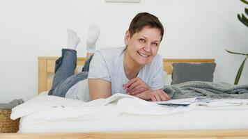 Relaxed senior woman lying on bed and reading book indoors at home. Leisure activity of mature female at comfort modern flat. Casual elderly life and wrinkled skin closeup video