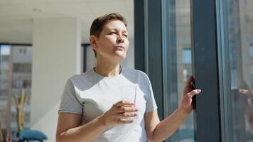 Sénior femme les boissons l'eau permanent près le fenêtre dans une lumière pièce video