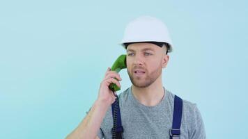 Serviceman in overalls and helmet speaks on an old telephone and consults a client on a blue background video