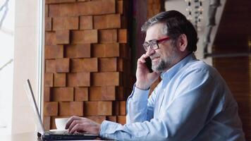 Close up of senior businessman talking on the mobile phone looking at laptop in cafe video