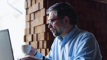 Senior freelancer businessman in glasses typing text on the keyboard in coffeshop and drink coffee video