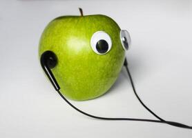 verde pequeño manzana con linda ojos y auriculares en blanco antecedentes. conceptual foto. foto