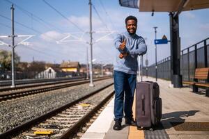 contento hombre con maleta en pie en ferrocarril estación y demostración pulgar arriba. foto