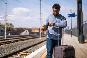 Tired man looking at clock and drinking coffee while standing with suitcase on a railway station. photo