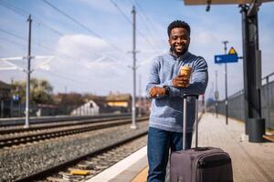 contento hombre con maleta disfruta Bebiendo café mientras en pie en ferrocarril estación. foto