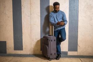 Happy man with suitcase using phone while leaning against a wall in passage of the railway station. Copy space on image for your advert or text. photo