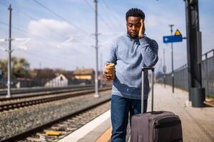 cansado hombre Bebiendo café mientras en pie con maleta en un ferrocarril estación. foto