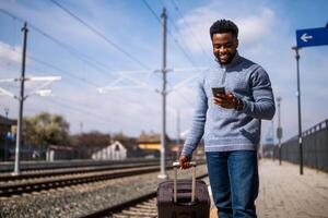 contento hombre con un maleta utilizando teléfono mientras en pie en el ferrocarril estación foto