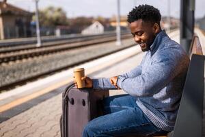 Happy man with suitcase and coffee looking a clock while sitting on a bench at the railway station. photo