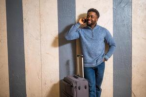 Happy man with suitcase using phone while leaning against a wall in passage of the railway station. Copy space on image for your advert or text. photo