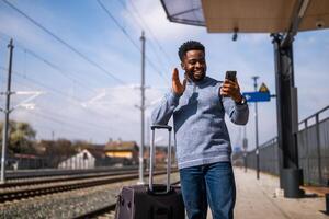 contento hombre con un maleta tomando selfie y ondulación en un ferrocarril estación. foto