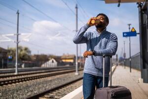 contento hombre con maleta disfruta Bebiendo café mientras en pie en ferrocarril estación. foto