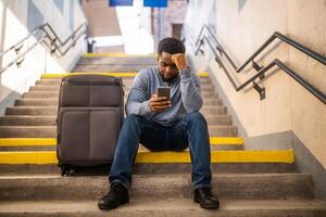 preocupado hombre con un teléfono y maleta sentado en un escalera a el ferrocarril estación. foto