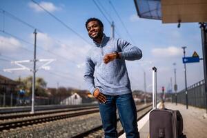 contento hombre mirando a su reloj mientras en pie con maleta en un ferrocarril estación. foto