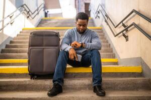 preocupado hombre mirando a reloj con un teléfono y maleta mientras sentado en un escalera a el ferrocarril estación. foto
