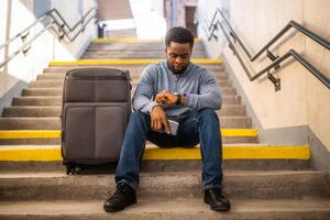 Worried man looking at clock with a phone and suitcase while sitting on a stairs at the railway station. photo