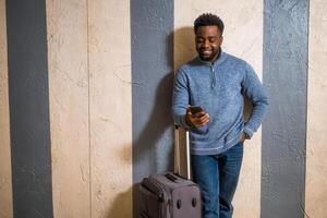 Happy man with suitcase using phone while leaning against a wall in passage of the railway station. Copy space on image for your advert or text. photo