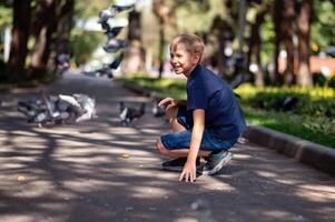 un chico en un azul camiseta se sienta en el asfalto en el parque y mira a el palomas foto