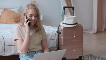 aberto fronteiras, pacote coisas, feliz férias, reserva hotel. feliz mulher falando em a telefone enquanto sentado em a chão Próximo para uma mala de viagem com coisas video