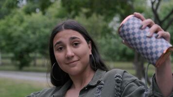 alegre hipster morena niña con largo pelo en verde ropa bailando en el parque a el música mientras participación un portátil altavoz en su mano. de cerca video