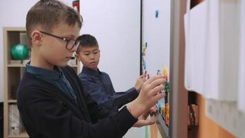 dos niños de diferente Razas competir a formar palabras en un magnético tablero durante un lección en clase.escuela para niños, enseñando adolescentes, ganancia conocimiento, aprender el idioma. video