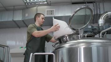 Worker A male brewer in uniform pours crushed malt into a beer tank for the production of craft beer. Close-up video