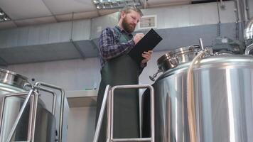 Adult Male brewer in an apron with a beard stands near the beer brewing tank and records the readings. Production of craft beer video