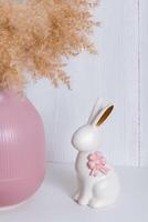 A white porcelain rabbit with a pink bow on a shelf next to a pink vase and a reed photo