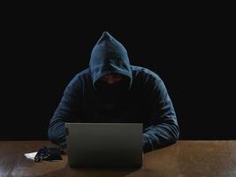 Portrait hacker spy man one person in black hoodie sitting on table looking computer laptop used login password attack security to data digital internet network system night dark background copy space photo