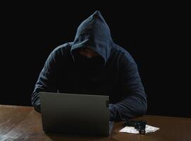 Portrait hacker spy man one person in black hoodie sitting on table looking computer laptop used login password attack security to data digital internet network system night dark background copy space photo