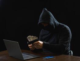 Portrait hacker spy man one person in black hoodie sitting on table looking computer laptop used login password attack security to data digital internet network system night dark background copy space photo