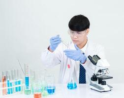 retrato asiático hombre estudiante científico vistiendo un médico vestido en el laboratorio mirando mano a químico. causado por mezcla reactivos en científico investigación laboratorios con prueba tubos y microscopio en el mesa foto