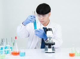 retrato asiático hombre estudiante científico vistiendo un médico vestido en el laboratorio mirando mano a químico. causado por mezcla reactivos en científico investigación laboratorios con prueba tubos y microscopio en el mesa foto