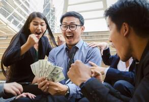 Team of young successful and excited business people sitting on steps outside office building. A man with eyeglasses holding dollar banknotes in hands photo