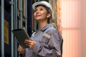 felicidad hembra ingeniero trabajando a internacional Envío carga patio trasero. mujer en uniforme utilizando digital tableta mientras mirando a envase para inspección cargando información foto