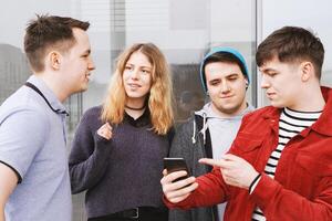 grupo de Adolescente amigos teniendo un conversacion mientras joven hombre es demostración alguna cosa en su teléfono inteligente foto