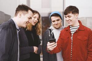 joven hombre demostración alguna cosa gracioso en su teléfono inteligente a un grupo de Adolescente amigos foto